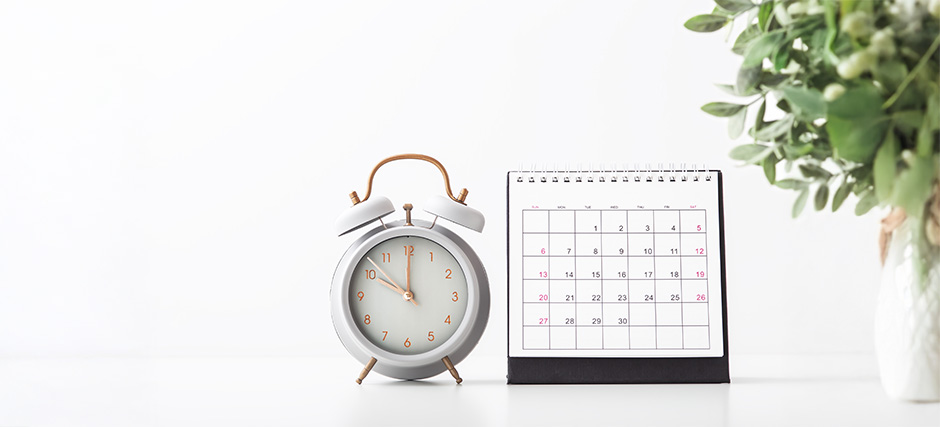 Minimalist setup with gold-handled white alarm clock showing 700, desk calendar with 31-day layout, and leafy potted plant on white background.