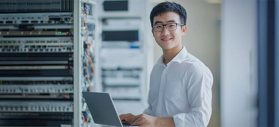 Person in white shirt using laptop in server room with racks of networking equipment and cables